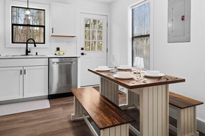 Une cuisine avec des meubles blancs, un lave-vaisselle en acier inoxydable, une table à manger en bois avec des bancs, et une fenêtre donnant sur les arbres.