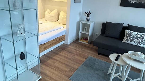 A modern living room with a bed, a sofa, a glass cabinet, and a dining area. - FeWo am Deich 2, modern apartment with balcony and elevator (Bremerhaven)