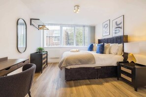 Ein modernes Schlafzimmer mit einem Bett, Nachttischen, einem Schreibtisch, einem Stuhl und einem Fenster mit Jalousien.