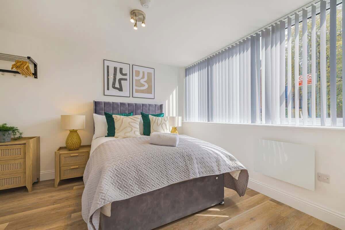 A modern bedroom with a large bed, a nightstand with a lamp, and a window with blinds.