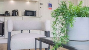 Una cocina moderna con armarios blancos, un microondas y una cafetera. Se observa una silla con cojín blanco y una planta en maceta al frente.