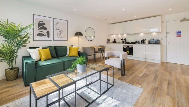 Una sala de estar moderna con un sofá verde, una mesa de centro y un comedor con una silla blanca. Se observa una cocina con armarios blancos y un microondas.