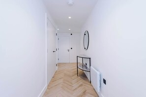 A hallway with white walls, a wooden herringbone floor, a door at the end, and a small table with a mirror on the wall. - Stylish 2 Bed 2 Bath in Central Manchester (Manchester)