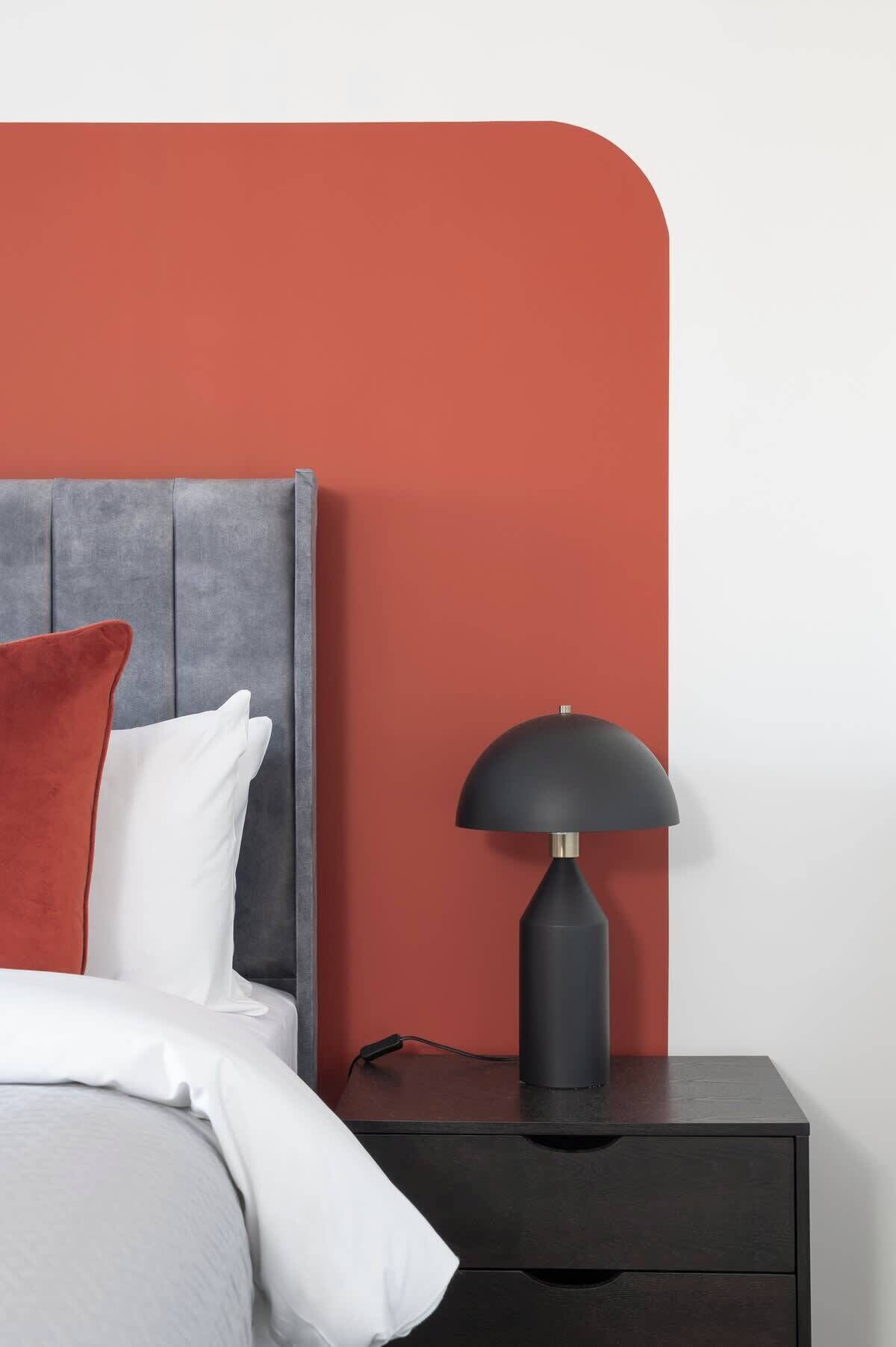 A bed with white and red pillows, a grey headboard, and a black nightstand with a lamp.