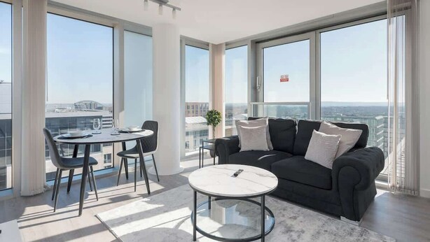 Ein modernes Wohnzimmer mit großem Fenster, einem runden Glastisch, einer dunklen Couch und einem Essbereich mit Stühlen und Tisch.