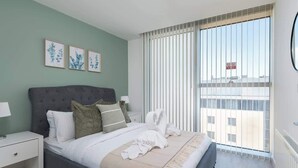 Ein Schlafzimmer mit einem Bett, einem Nachttisch, einem Fenster mit Jalousien und gerahmten Pflanzenbildern an der Wand.