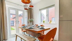 Ett modernt matområde med ett träbord och orange stolar, en bänk och ett fönster med utsikt över en byggnad med tegelstensfasad.