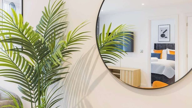Habitació d'hotel moderna amb un mirall rodó, un llit amb roba de llit taronja i blava, un armari de fusta i una planta gran en test.