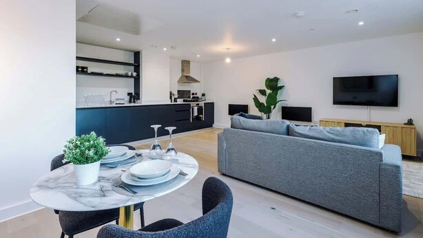A modern living room with a grey sofa, a dining table set with plates and glasses, a TV mounted on the wall, and a kitchen area in the background. - Stylish Manchester Apartment: City Access & Wi-Fi (Manchester)