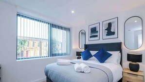 A modern hotel room with a large bed, a nightstand, a bedside table, and a window with blinds. - Stylish Manchester Apartment: City Access & Wi-Fi (Manchester)