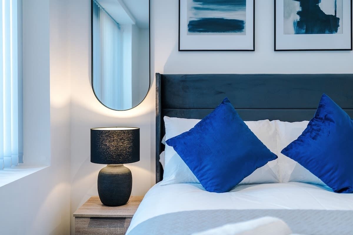 Ein ordentlich bezogenes Bett mit blauen Kissen, einer Nachttischlampe und einem gerahmten Kunstwerk darüber.