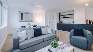 A modern living room with a grey sofa, a blue armchair, and a marble coffee table. There is a kitchen area with dark cabinets and a white countertop. - Stylish Manchester Apartment: City Access & Wi-Fi (Manchester)