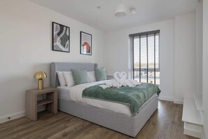 A modern bedroom with a bed, a nightstand, a window with blinds, and two framed artworks on the wall. - Modern Canal-Side Apartment in Milton Keynes (Milton Keynes)