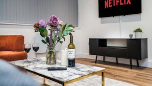 Ein Wohnzimmer mit einem Marmortisch, einer Fernbedienung, einer Weinflasche und einem Blumenstrauß. Eine schwarze TV-Konsole mit dem Schriftzug 'NETFLIX' ist an der Wand befestigt.