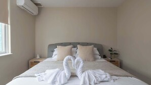 A hotel room with a bed, two bedside tables, a nightstand, a potted plant, and an air conditioning unit. - Better Than a Hotel | Parking | Stony Stratford (Milton Keynes)