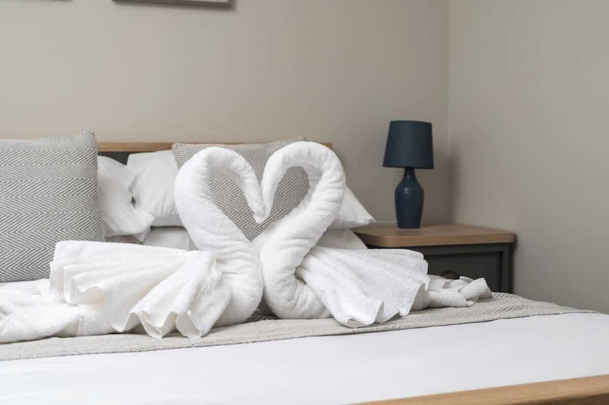 Een keurig opgemaakt bed met een handdoekje in hartvorm, een bedlampje en een gestreept kussen.