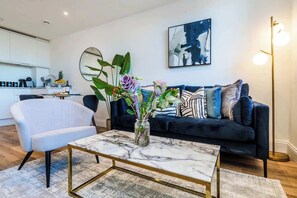 A modern living room with a dark blue sofa, a white armchair, and a marble coffee table. There is a vase with flowers on the coffee table. - Central Gravesend 1BR | Kitchen +Wi-Fi | Sleeps 2 (Gravesend)
