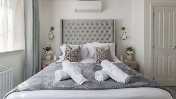 Cama bem arrumada com manta cinza e duas toalhas enroladas, cabeceira estofada, mesas de cabeceira com vasos decorativos e um aparelho de ar condicionado na parede.