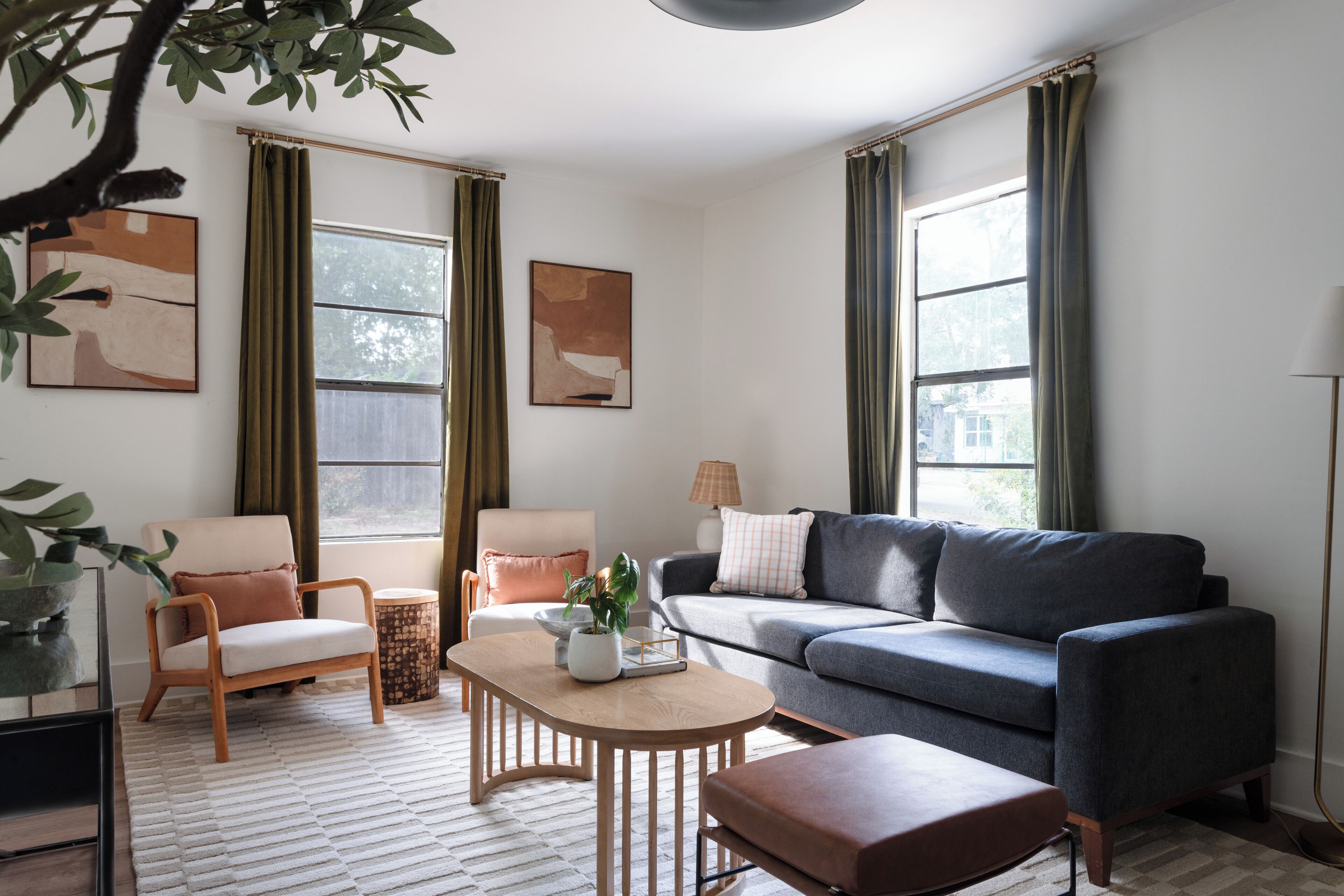 Una sala de estar moderna con un sofá gris, una mesa de centro de madera y un sillón blanco. Hay dos ventanas con cortinas verdes, una lámpara de pie y pinturas abstractas en las paredes.