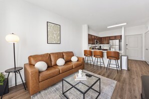 Ein modernes Wohnzimmer mit einem braunen Ledersofa, einem Glastisch und einer Stehlampe. Im Hintergrund ist eine Küche mit Holzausbau und Barhocker erkennbar.