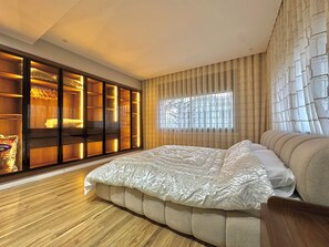 Une chambre à coucher avec un grand lit, un plancher en bois, une armoire avec des étagères éclairées et une fenêtre ornée de rideaux à motifs.