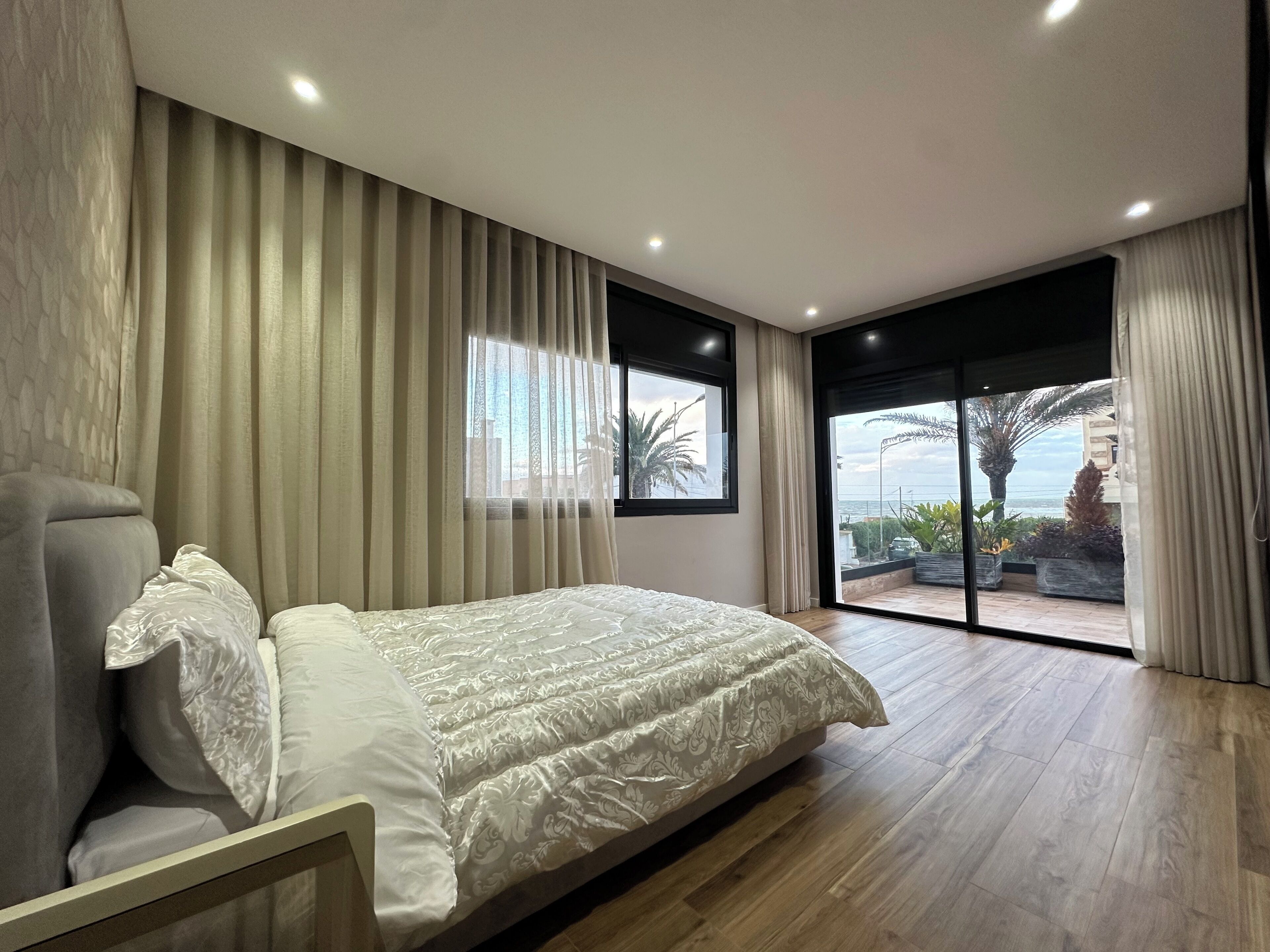A modern bedroom with a large bed, a view of the outdoors through a sliding glass door, and a balcony with plants.