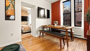 A modern dining area with a wooden table and bench, a flat-screen TV mounted on the wall, and a large window with red curtains. - NEW - Stylish Art Deco Apt • Easy Access to Manhattan (Hoboken)