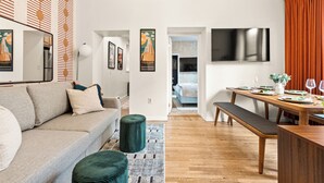 A modern living room with a sofa, a dining table, a bench, and a TV mounted on the wall. - NEW - Stylish Art Deco Apt • Easy Access to Manhattan (Hoboken)