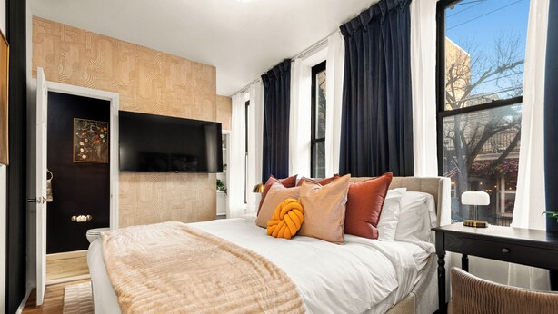 A modern hotel room with a large bed, a flat-screen TV mounted on the wall, and a view of the city through the window. - NEW - Stylish Art Deco Apt • Easy Access to Manhattan (Hoboken)