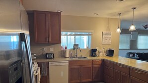 A kitchen with wooden cabinets, a dishwasher, a refrigerator, a window with blinds, and a coffee maker. - Hit the Trail or Lake: Spacious 4BR/2BA Retreat with Pool Table & Large Bar Area (Needles)