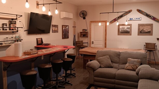 A cozy living room with a sofa, a coffee table, and a bar area with stools. There are framed pictures on the walls and a television mounted above the bar. - Hit the Trail or Lake: Spacious 4BR/2BA Retreat with Pool Table & Large Bar Area (Needles)