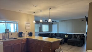 A modern kitchen with wooden cabinets, a large island, pendant lighting, and a view to a living area with a black sofa and patterned cushions. - Hit the Trail or Lake: Spacious 4BR/2BA Retreat with Pool Table & Large Bar Area (Needles)