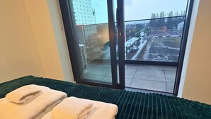Ett hotellrum med en säng, ett grönt, mönstrat sängöverkast och utsikt över en stad genom ett stort fönster.
