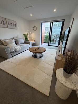 Een moderne woonkamer met een grijze bank, een ronde salontafel, een groot raam met uitzicht op een gebouw, en een klok aan de muur.