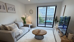 Ett modernt vardagsrum med en grå soffa, ett runt soffbord, ett stort fönster med utsikt över en byggnad och en platt-TV.