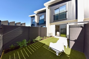 A modern building with a flat roof, large windows, and a fenced backyard with artificial grass and a white chair. - Stylish 2BR Townhouse with Balcony (Christchurch)