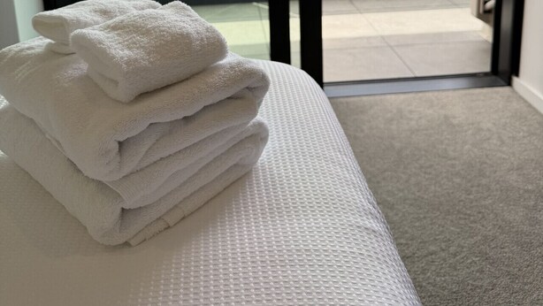 Ein Hotelzimmer mit einem Bett, einem Stapel weißer Handtücher und Blick auf ein Gebäude sowie einen Parkplatz durch eine Glastür.