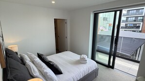 Ein modernes Hotelzimmer mit einem großen Bett, einem Nachttisch mit Lampe, einer Schiebeglastür zum Balkon und Blick auf die Stadtlandschaft.
