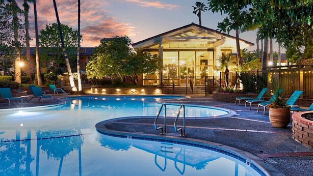 A swimming pool with lounge chairs and a glass-enclosed building in the background. - Disneyland Days, Surf City Waves, Newport Nights (Costa Mesa)