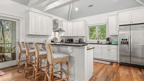 A kitchen with white cabinets, a stainless steel refrigerator, wicker bar stools, and a large window. - Pet friendly home located in the middle on Bald Head Island (Bald Head Island)