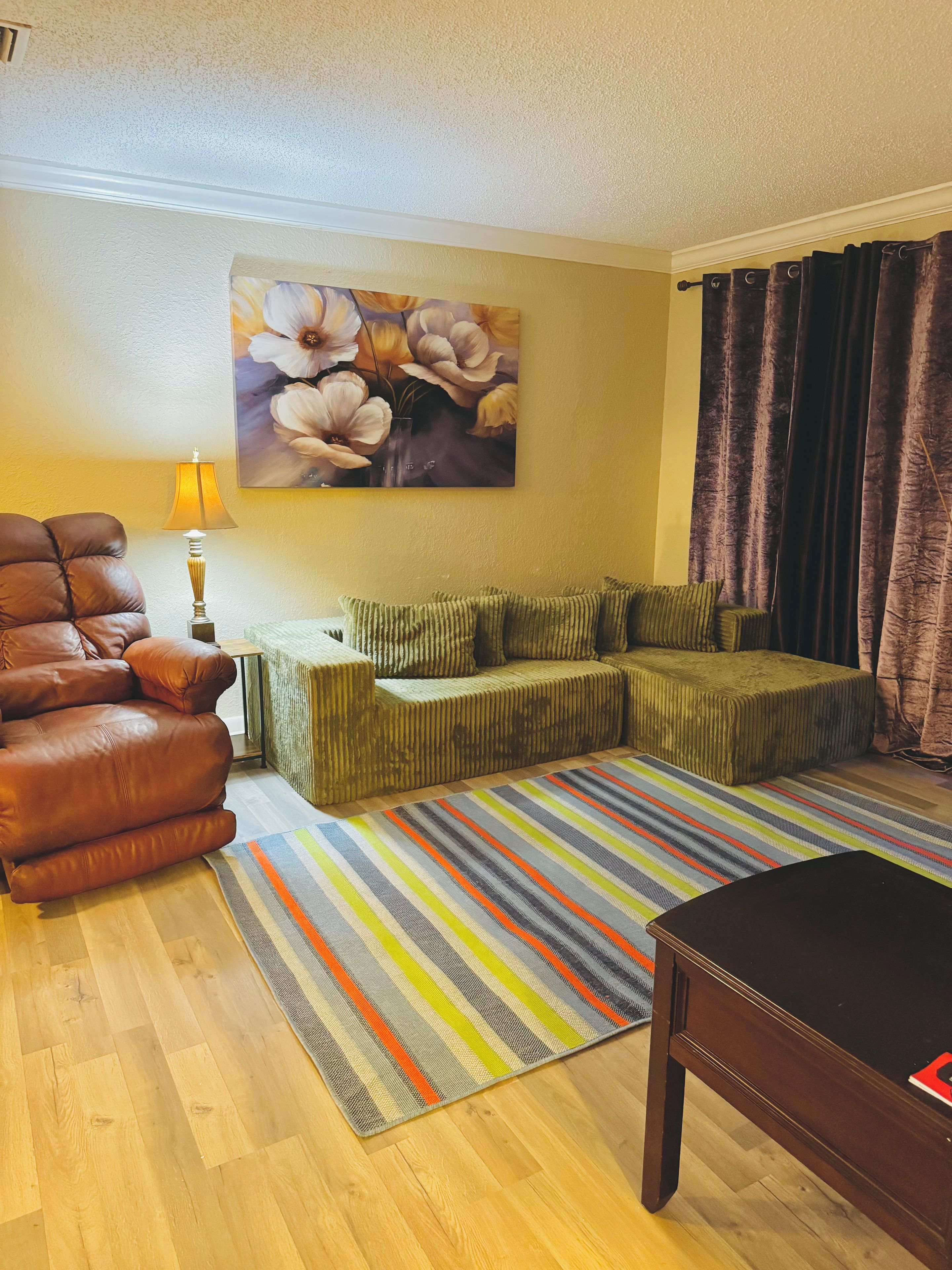 A living room with a leather sofa, a green sofa set, a striped rug, and a painting of flowers on the wall.