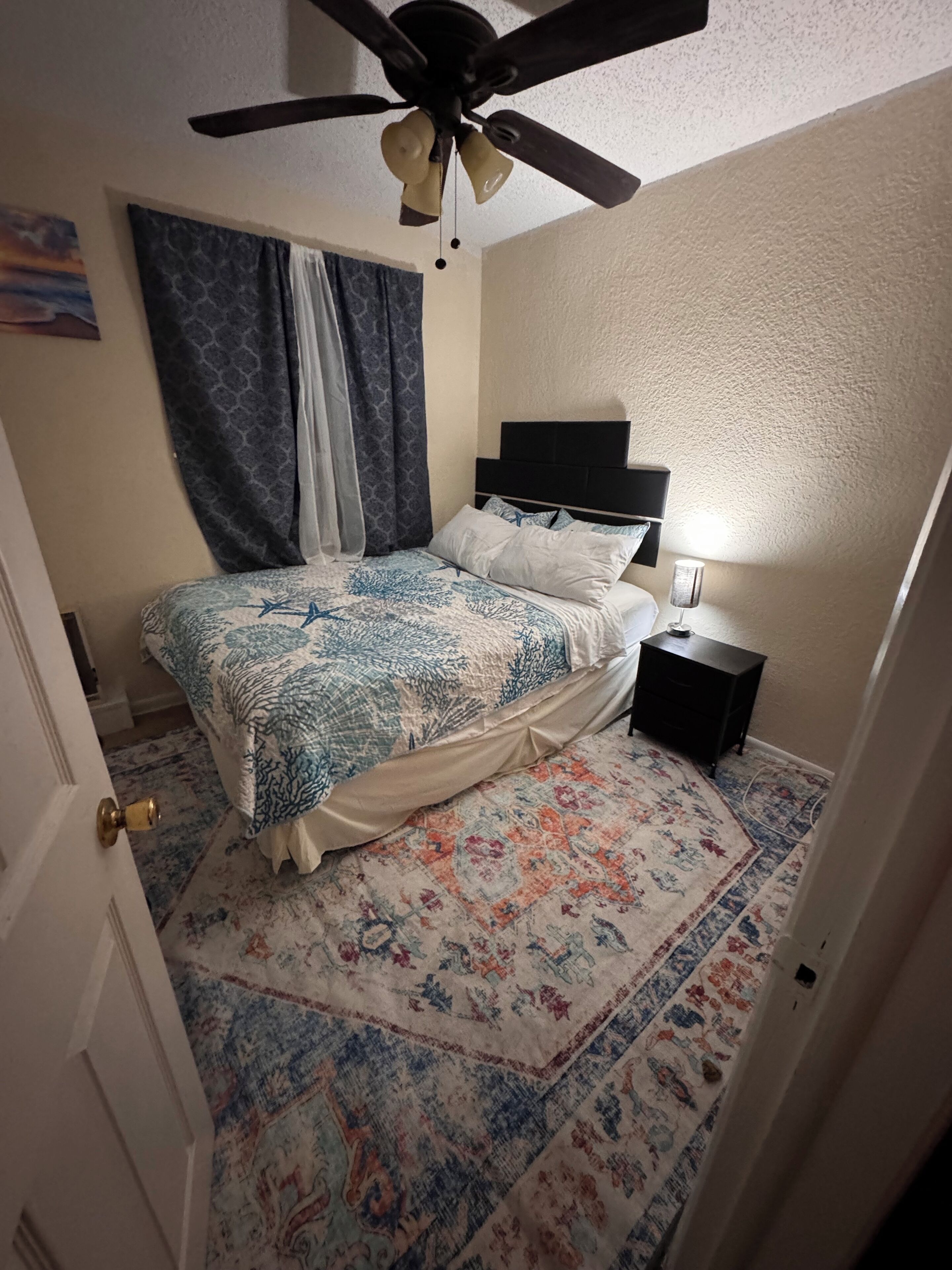 Un dormitorio con cama, ventilador de techo, mesita de noche, lámpara y alfombra estampada.