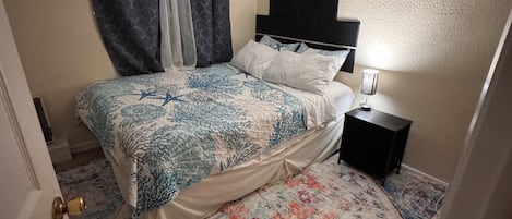Un dormitorio con cama, ventilador de techo, mesita de noche, lámpara y alfombra estampada.