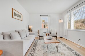 Ein modernes Wohnzimmer mit einer Couch, Sesseln, einem Couchtisch und einem Teppich. An der Wand hängt ein Schild mit der Aufschrift 'HOME', es steht eine Stehlampe und ein Fenster mit Jalousien ist zu sehen.