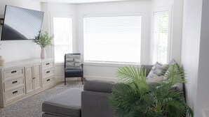 Un salon moderne avec un canapé gris, un téléviseur à écran plat fixé au mur et une grande fenêtre équipée de stores.
