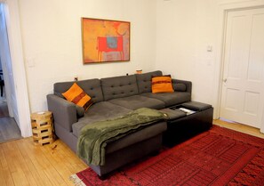 Ein Wohnzimmer mit einem grauen Ecksofa, einem roten Teppich mit Muster, einer grünen Decke und einem Bild an der Wand.