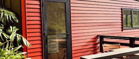 A red wooden building with a black door and windows, a deck with wooden furniture, and tropical plants.