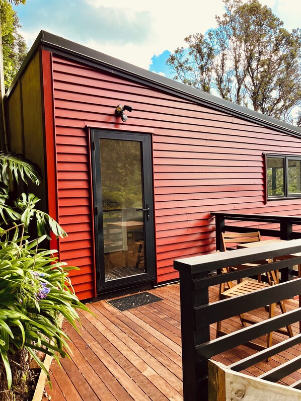 Ein rotes Holzgebäude mit schwarzem Tür- und Fensterrahmen, einer Terrasse mit Holzmöbeln und tropischen Pflanzen.