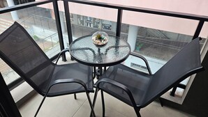 Un balcón acristalado con una mesa redonda y dos sillas, una pequeña planta en maceta, y vista a la calle.