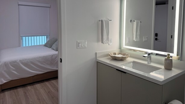 Une salle de bain moderne avec un grand miroir, un lavabo et des toilettes. La pièce adjacente contient un lit avec du linge de lit blanc.
